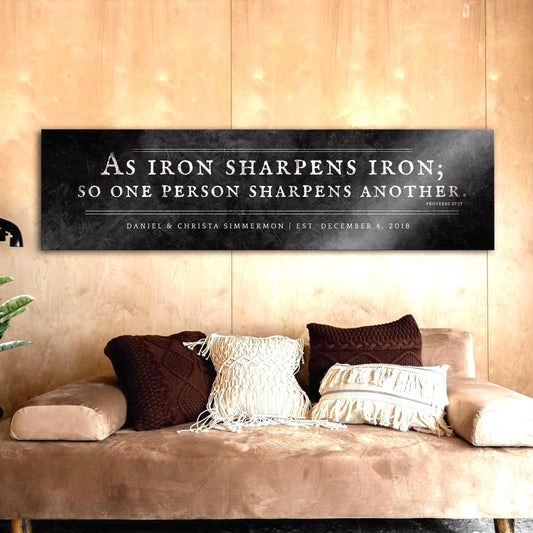 Distressed Iron Alloy As Iron Sharpens Iron Sign This wide format wall decor features the words of Proverbs 27:17inscribed in silver on a distressed dark wrought iron finish with an option to add a short personalization or dedication beneath the scripture. The sign is wall mounted above a sofa and is personalized with the names of a married couple and their union date. Photo courtesy of Honeycomb Proverbs