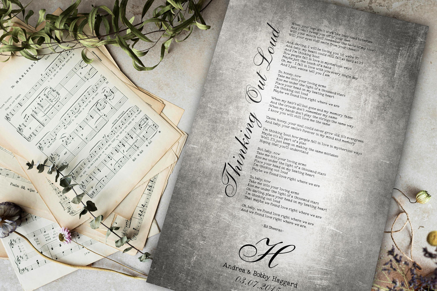 A vintage style tin wedding song plaque, laying on a tabletop next to an aged stack of sheet music and lightly accented by sprigs of greenery. The plaque is crafted on tarnished tin, the traditional anniversary gift metal for the 10th wedding anniversary, and personalized with first dance song lyrics, the couple's wedding date, names, and last name monogram letter.