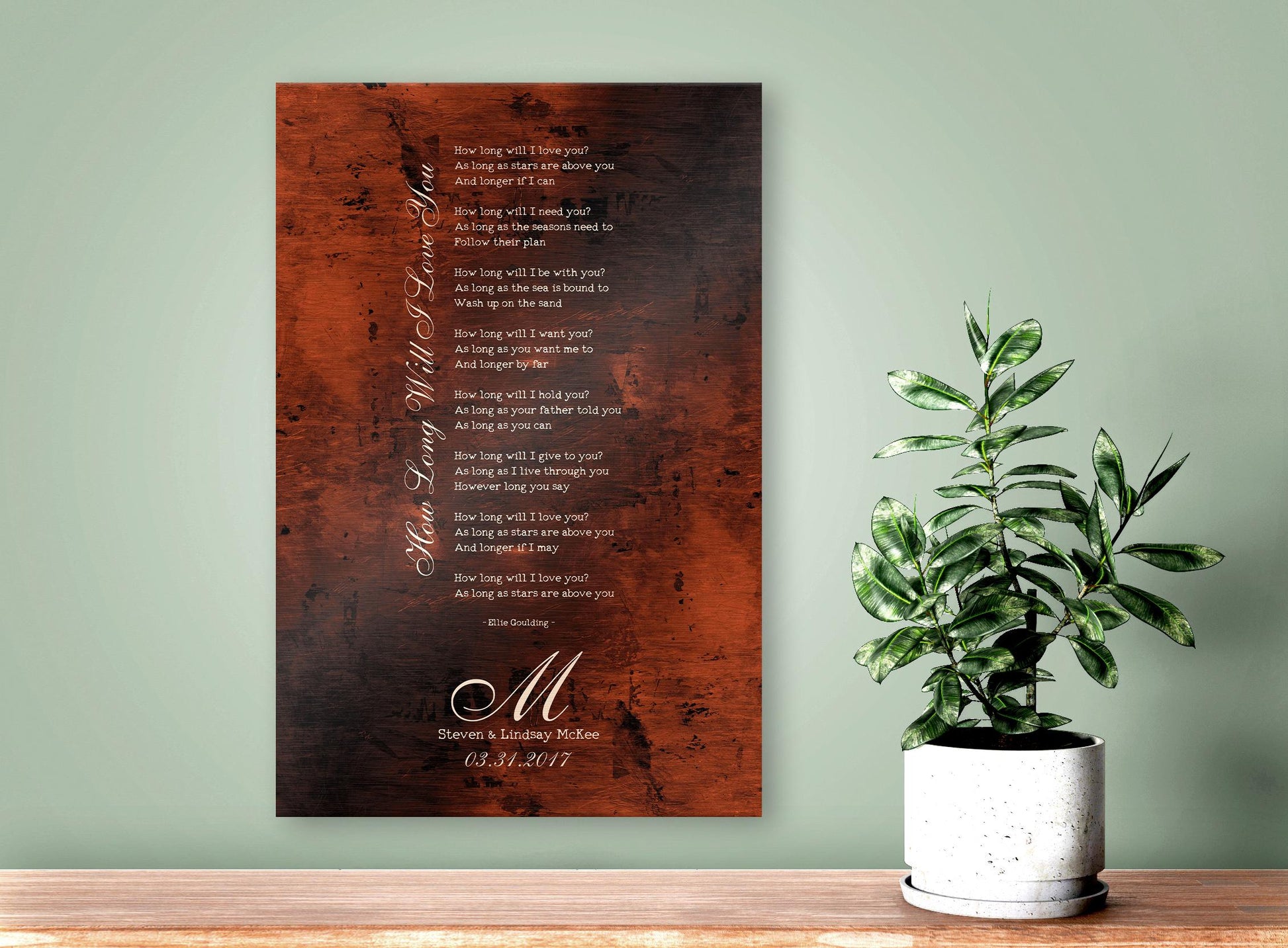 A burnished bronze anniversary gift plaque is personalized with a calligraphic monogram letter,  the wedding anniversary date and names of a married couple. Above is their special wedding song, laid out in a traditional lyrical stanza with the song title elegantly placed vertically, along the left margin. The wedding song lyric art is wall hung on a sage green wall above a shelf where a potted plant also accents the artwork on the right hand side of the frame.