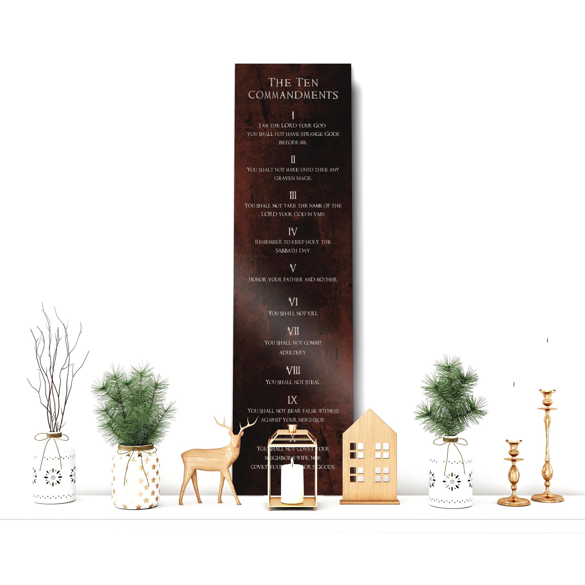 May include: A rustic rubbed bronze metal sign with the Ten Commandments written in white text. The sign is hanging on a white wall with a decorative shelf below it. The shelf has a white vase with a green plant, a white vase with a brown plant, a gold candle holder with a white candle, a gold candle holder with a white candle, a wooden house decoration, and a gold deer figurine.