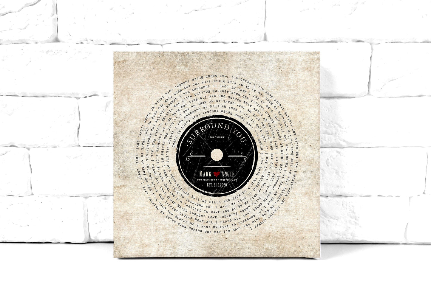 lyrics on canvas, custom song art with lyrics set in a vinyl record design around a retro record label personalized with a couple's first names, with a heart stamp, and the song name and performing artist of choice. Art also features the statement "2 years down, forever to go" as a nod to the 2nd wedding anniversary. This cotton canvas song art is leaning against a white brick wall.