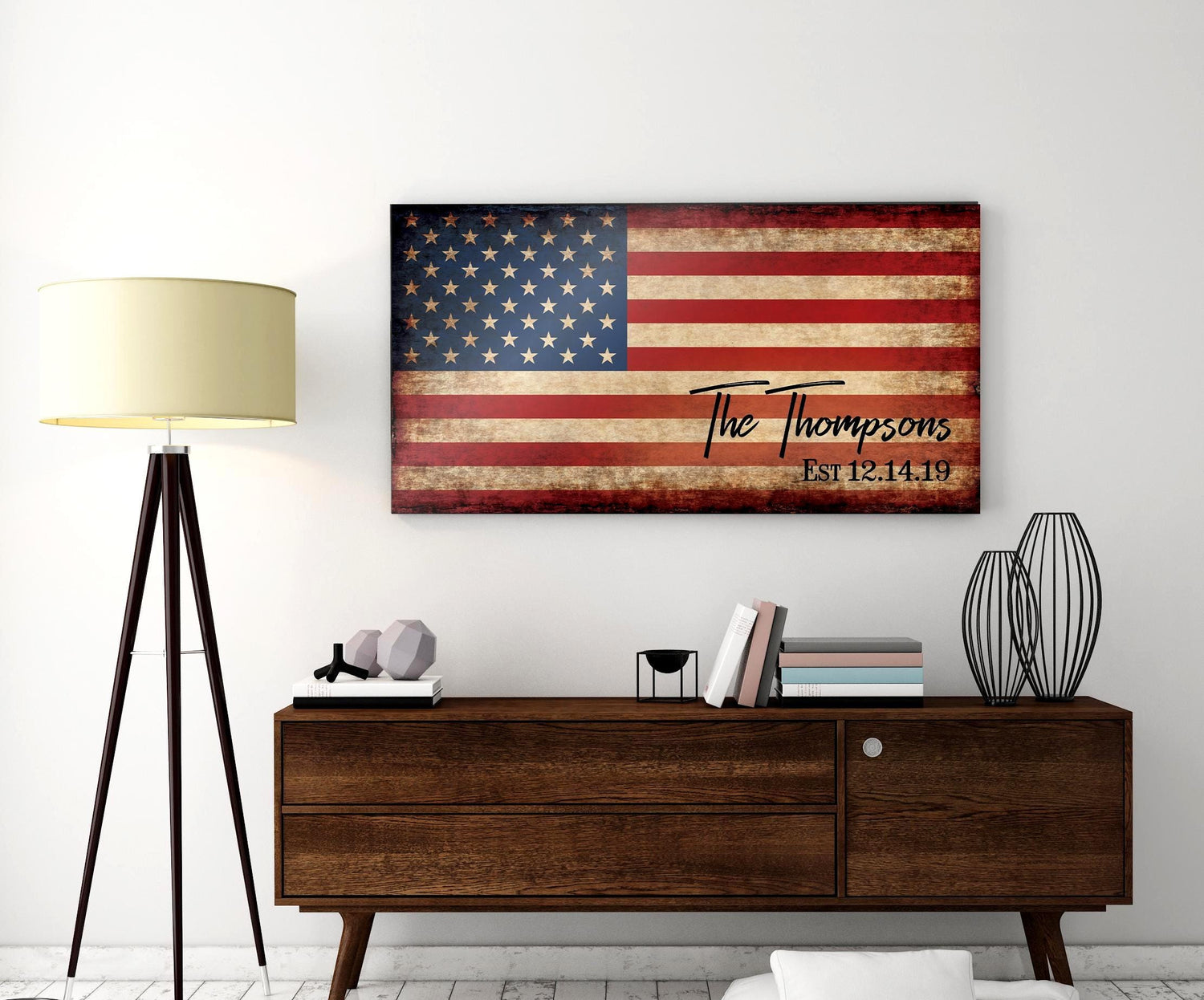 A distressed wood sign with an American flag design and the text "The Thompsons Est 12.14.19". The sign is hanging on a white wall above a wooden dresser.