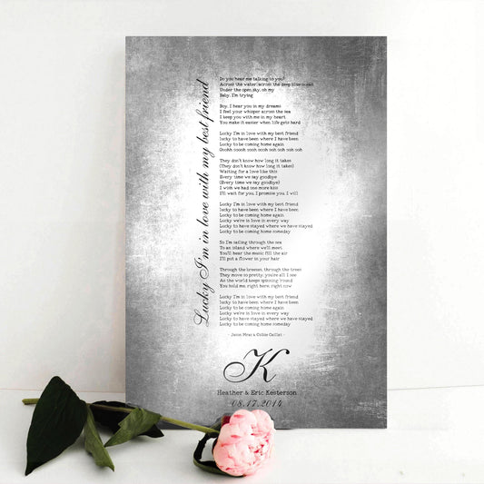 Couples custom song lyrics wall decor - vintage metal tin aluminum song plaque sits on a white background with a single pink rose. The sign features the inscription “lucky I’m in love with my best friend” in an elegant calligraphy running vertically up the side of song lyrics in a stanza format - which is an except from the lyrics. Beneath is a last name Monogram letter, names and a union date, for posterity.  Photo courtesy of Honeycomb Proverbs