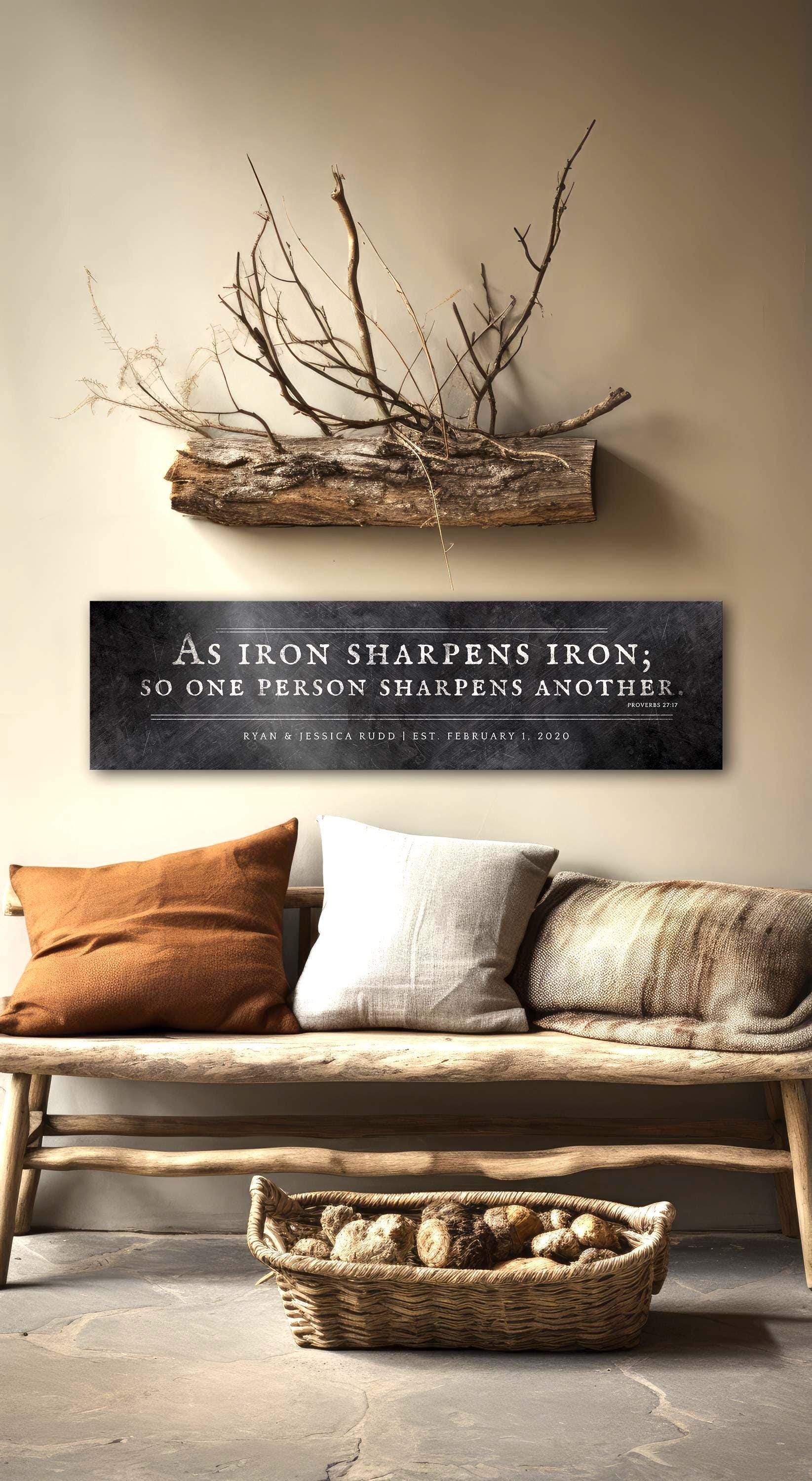 Distressed Iron Alloy As Iron Sharpens Iron Sign This wide format wall decor features the words of Proverbs 27:17inscribed in silver on a distressed dark wrought iron finish with an option to add a short personalization or dedication beneath the scripture. The sign is wall mounted above a sofa and is personalized with the names of a married couple and their union date. Photo courtesy of Honeycomb Proverbs
