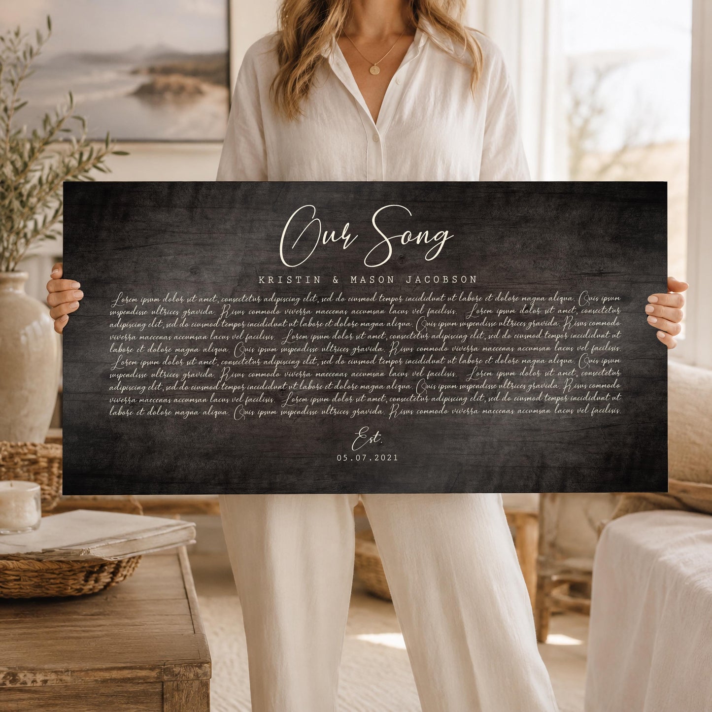 A large, horizontal and personalized song plaque on a barnwood in a dark grey tone with song lyrics is held by a blond woman wearing cream top and pants.