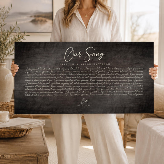 A large, horizontal and personalized song plaque on a barnwood in a dark grey tone with song lyrics is held by a blond woman wearing cream top and pants.