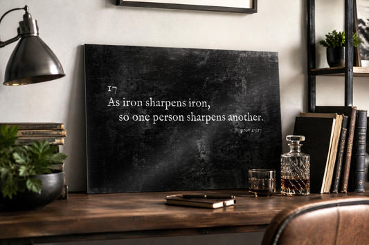 Industrial style Christian metal plaque featuring As iron sharpens iron, so one person sharpens another of the Bible's Proverbs 27:17. The plaque is leaning against a wall on a desktop of a man's desk next to books and a plant.